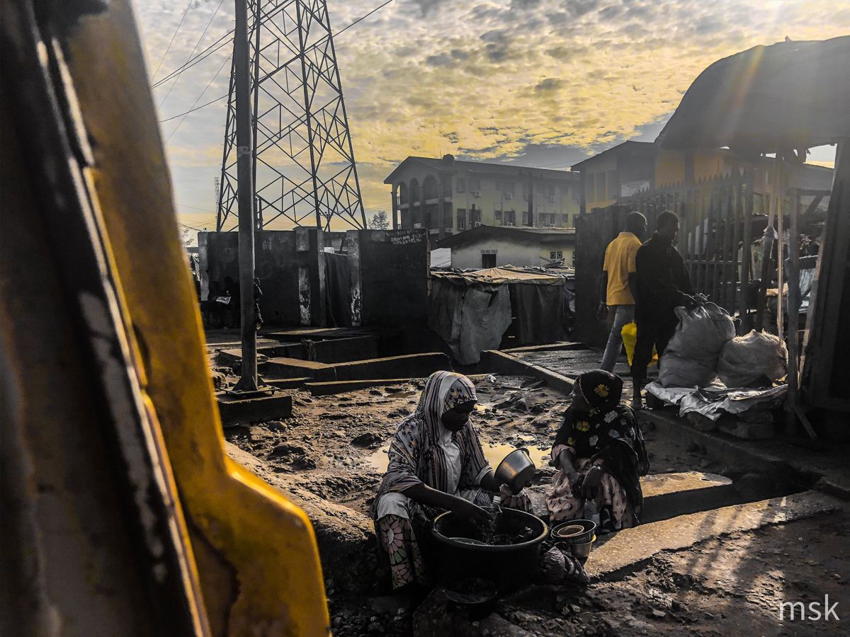Women washing bowls, framed through a yellow danfo window at sunrise